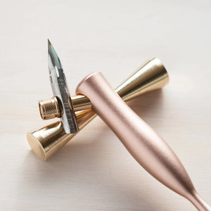 Tom's Studio Butterfly brass pen rest holding a rose gold calligraphy pen, on pale wood background